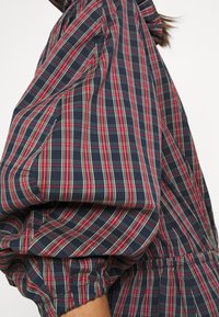 Marinblå rutiga skjorta med djärva röda och gröna linjer, puffärmar och elastisk midja för en texturerad, lager-på-lager look.