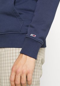 Navy blue hoodie made of cotton blend fabric, featuring ribbed cuffs and a small logo patch with red and white accents on the sleeve.