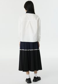 Camicia bianca a maniche lunghe con colletto nero, abbinata a una gonna plissettata con sezioni blu navy e nere, ulteriormente arricchita da un bordo in pizzo bianco.
