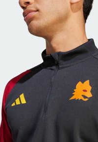 Sudadera negra deportiva con cremallera de un cuarto y mangas rojas, que presenta un logo de tres rayas amarillas y un gráfico de la cabeza de un lobo naranja en el pecho.