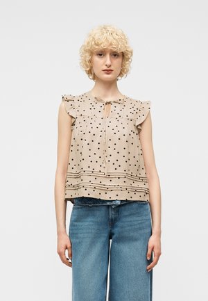 Jeune personne aux cheveux blonds bouclés portant un chemisier beige à pois sans manches avec des volants et un jean bleu, debout devant un fond uni.