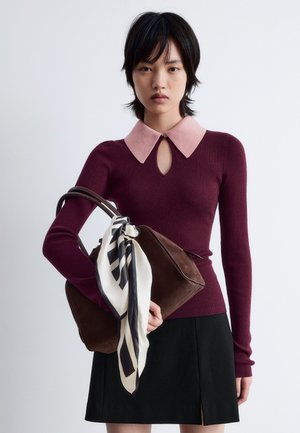 Burgunderfarbener gerippter Pullover mit einem rosafarbenen Kragen und einem Schlüsselloch-Detail, kombiniert mit einer braunen Handtasche, die mit einem weiß-schwarz gemusterten Schal akzentuiert ist.