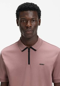 Polo a maniche corte in tessuto rosa chiaro con collo nero a zip e piccolo logo "HUGO" nero sul petto. Tessuto dalla texture liscia.