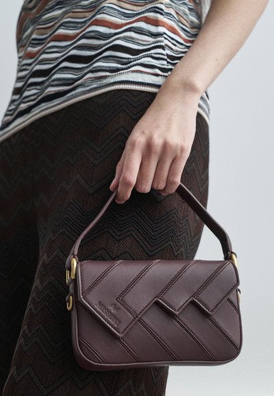 Hand som håller en liten mörkbrun läderhandväska med diagonalt stygn, guldfärgad hårdvara, bärs med mönstrade bruna byxor och randig topp.