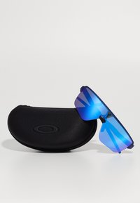 Gafas de sol con grandes lentes espejo azules, un marco rectangular y acentos en negro, colocadas junto a un estuche protector negro.