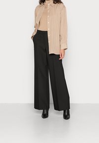 Kobieta w beżowym golfie, w beżowo-białą w paski koszulę z guzikami, czarnych szerokich spodniach i czarnych botkach, stojąca na białym tle.