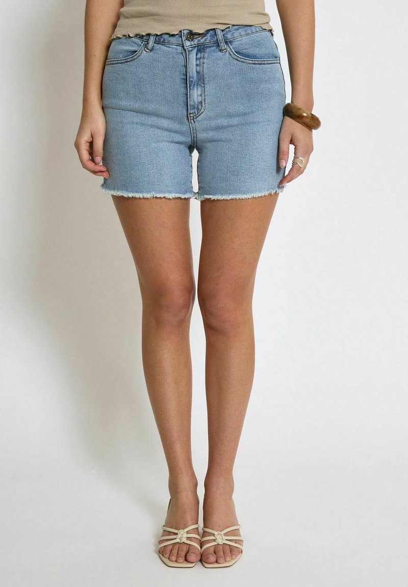 Shorts de mezclilla en un tono azul claro con dobladillo deshilachado, cintura alta y cinco bolsillos. Combinados con sandalias beige de tiras.