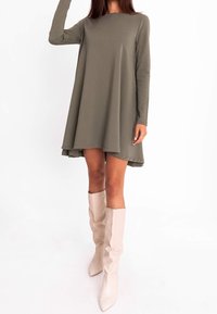 Olivgrünes, langärmeliges Kleid mit lockerem Sitz, rundem Ausschnitt und einem hinten tiefer geschnittenen Saum. Kombiniert mit beigefarbenen kniehohen Stiefeln.