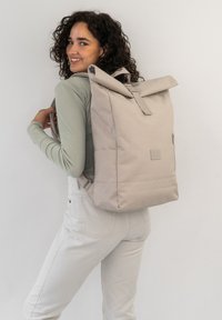 Roll-top backpack in light beige fabric with adjustable straps, a front zip pocket, and subtle branding. Smooth texture and minimalist design.