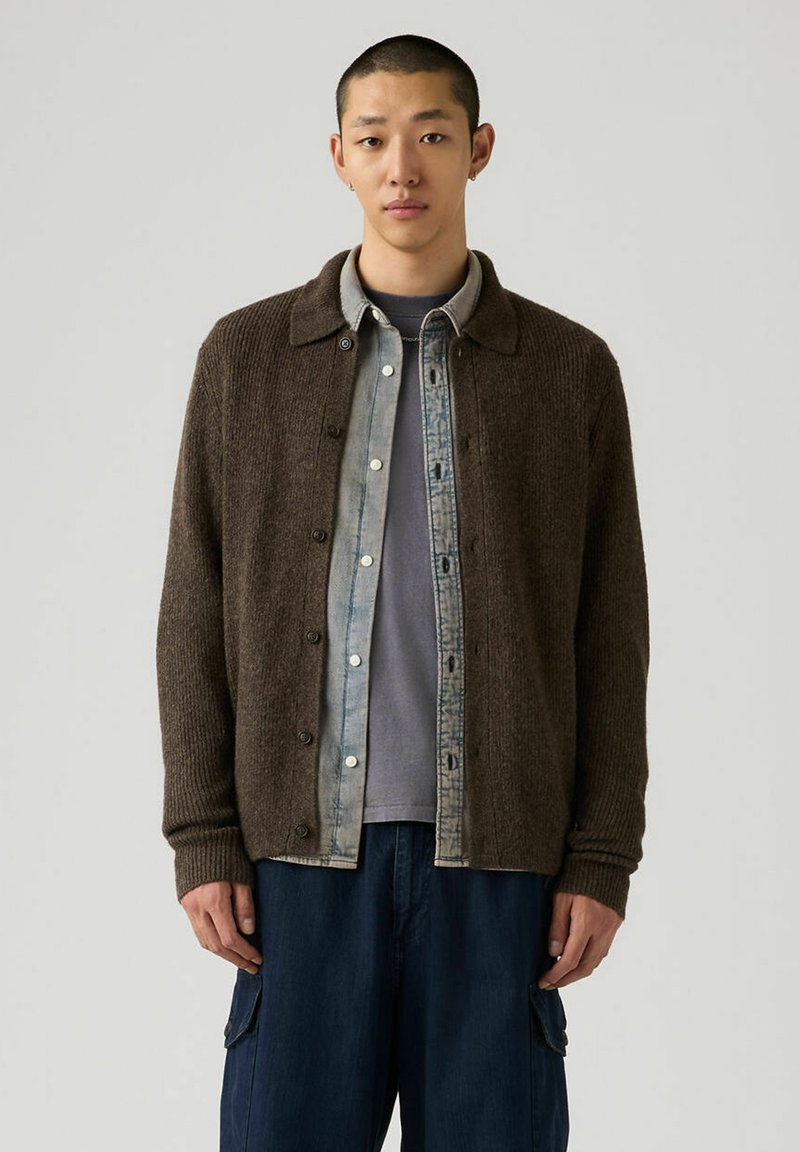 Cardigan en maille côtelée marron avec fermeture à boutons, porté sur une chemise en denim bleu clair et un t-shirt gris, associé à un pantalon décontracté bleu foncé.