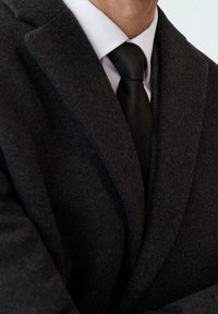 Donkergrijs wollen overjas met een geknipte kraag, losse pasvorm, gelaagd over een wit overhemd en een gestructureerde zwarte stropdas.