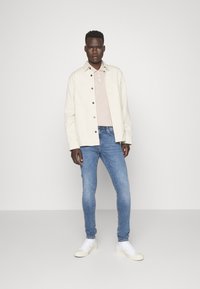 Chaqueta blanca, polo de canalé marrón claro, jeans ajustados azules y zapatillas blancas. La chaqueta tiene botones oscuros; el conjunto presenta un diseño casual.