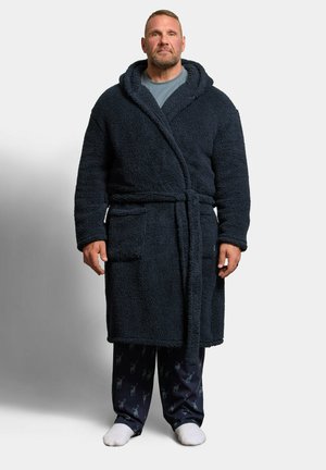 Un homme portant une robe polaire bleu foncé par-dessus une chemise grise et un pantalon de pyjama à motifs, debout avec des chaussettes blanches sur un fond neutre.