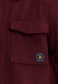 Bordová vlnená bunda s prednou kapsou a označením značky so žltým dizajnom. Mäkká textura s pevnou siluetou.