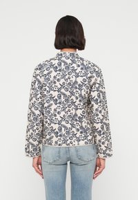 Μπουφάν με λουλουδάτο σχέδιο σε κρεμ χρώμα με μπλε σκούρα σχέδια, με μακριά μανίκια και στρογγυλεμένο γιακά, συνδυασμένο με ανοιχτό μπλε τζιν.