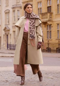 Trench coat bej cu guler larg, bluză roz, pantaloni largi maro și eșarfă cu imprimeu leopard, asortate cu mănuși maro și cizme cu toc strălucitoare.