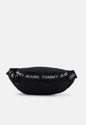 Zwarte stoffen heuptas met een gebogen vorm, voorzien van een wit tekstlogo "TOMMY JEANS" langs de band en een klein label met rode en blauwe accenten.