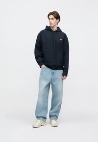 Μπλε ναυτικό φούτερ με κουκούλα και μπροστινή τσέπη, με κόκκινο λογότυπο, συνδυασμένο με ανοιχτό μπλε φαρδιά τζιν και μπεζ αθλητικά παπούτσια.