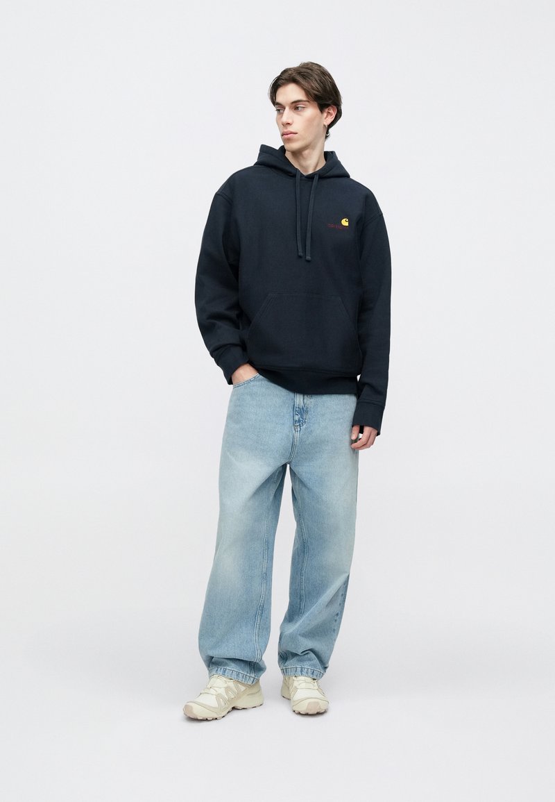 Marineblauwe capuchontrui met een zak aan de voorkant en een rood logo, gecombineerd met lichtblauwe losse spijkerbroek en beige sneakers.