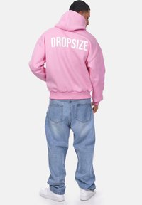 Felpa rosa con testo "DROPSIZE" sulla schiena, abbinata a jeans larghi di un azzurro chiaro e scarpe da ginnastica bianche. Tessuto morbido, vestibilità casual.