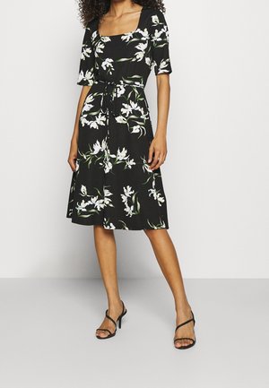 Schwarzes Kleid mit kurzen Ärmeln, quadratischem Ausschnitt, floralem weißen und grünen Muster, tailliert mit Band und A-Linien-Rock. Schwarze Riemchen-Heels.