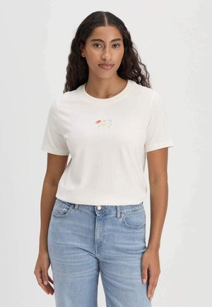 Vrouw met lang krullend haar, gekleed in een wit t-shirt met kleine geborduurde bloemen en lichtblauwe spijkerbroek, staand tegen een effen achtergrond.