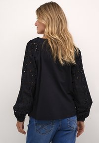 Schwarzes Oberteil mit bestickten Lochdetails an den Ärmeln, lockerer Schnitt und Knopfmanschetten, kombiniert mit Jeans.