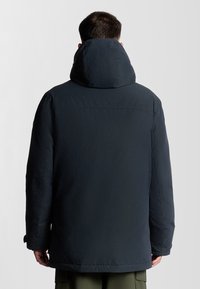 Mörkblå pufferjacka med hög krage och huva. Har en slät yta och avslappnad passform. Ärmsluten har justerbara remmar.