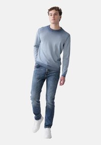 Hellblauer Rundhalsausschnitt-Pullover mit langen Ärmeln, kombiniert mit dunkelblauen Jeans und weißen Turnschuhen, was einen lässigen, gestuften Look ergibt.