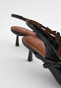 Slingback heels in black patent leather with a brown insole, mid-height block heel, and decorative bow accent on the strap.