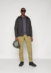 Zwarte lichtgewicht jas over een grijs shirt, gecombineerd met beige cargobroek en zwarte sneakers. Houdt een grijze fietshelm vast.