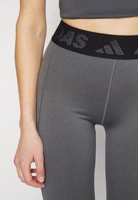 Leggings sportivi grigi con una cintura elastica nera che presenta un logo ripetuto. Il tessuto ha una texture liscia e elastica e le cuciture sono visibili.