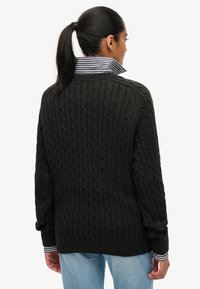 Zwarte kabelgebreide trui met een gestructureerd patroon, voorzien van een contrasterende gestreepte kraag en een ontspannen pasvorm, gezien vanaf de achterkant.