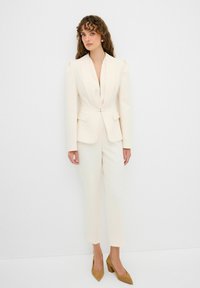 Crème-kleurige slim fit blazer met pieklapels en aangepaste broek. Gladde stof, gestructureerde schouders en tan gekleurde suède hakken maken de look compleet.