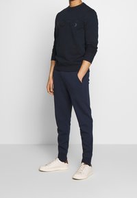 Uomo con felpa blu navy e pantaloni sportivi con sneakers bianche, in piedi con una mano in tasca davanti a uno sfondo neutro.