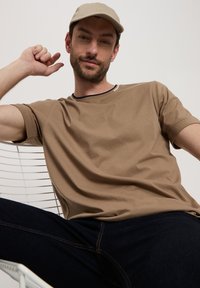 Brązowy t-shirt z krótkim rękawem i okrągłym dekoltem, który ma kontrastowe wykończenie. Model siedzi, nosząc beżową czapkę i ciemne dżinsy.