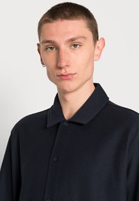 Camisa azul-marinho de botões com colarinho, apresentando um tecido texturizado e dois botões na frente, design estruturado sem padrões ou acentos.