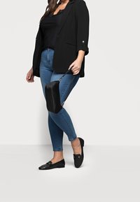Zwarte blazer, zwarte kanten top, blauwe denim jeans, zwarte loafers met gouden accenten en een kleine zwarte crossbody tas. Casual outfit presentatie.
