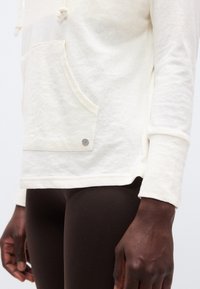 Κοντινό πλάνο ενός ατόμου που φοράει μπεζ φούτερ με κουκούλα και μπροστινή τσέπη, καθώς και σκούρο καφέ εφαρμοστά παντελόνια, μπροστά σε απλό φόντο.