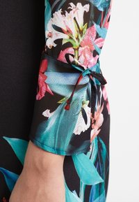 Blommönstrad klänning med livfulla rosa, gröna och teal-färger. Har en texturerad tygkvalitet och ett knyt detaljer vid ärmen.