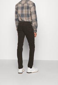 Pantalon en velours côtelé brun foncé à coupe slim, associé à une chemise à carreaux aux couleurs sobres et des baskets blanches. Le tissu a une surface texturée.