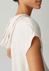 Kremowy top z krótkim rękawem z kapturem. Wykonany z gładkiego, lekkiego materiału. Charakteryzuje się luźnym krojem i subtelnymi detalami szwów.