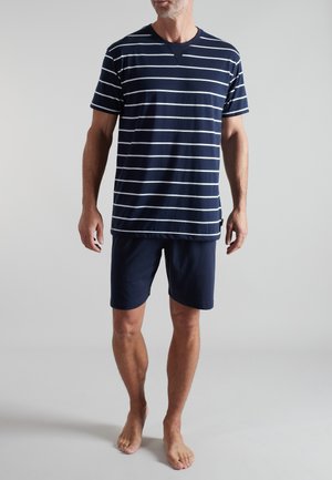 Mann trägt marineblaue Shorts und ein marineblaues T-Shirt mit weißen horizontalen Streifen, steht barfuß vor einfachem Hintergrund.