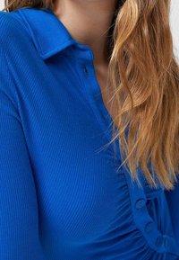 Haut bleu côtelé avec col, doté d'une fermeture à bouton et de détails froncés sur un côté, présentant une texture lisse et une couleur vive.