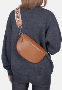 Braune Leder-Crossbody-Tasche mit runder Form, strukturiertem Oberflächenmaterial und einem gemusterten verstellbaren Trageriemen, die auf dem Rücken über einem dunklen Pullover getragen wird.