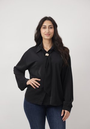 Vrouw met lang donker haar, gekleed in een zwarte blouse met een broche en een blauwe spijkerbroek, staand met één hand op haar heup tegen een effen achtergrond.