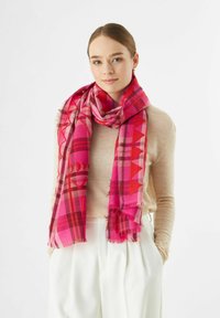 Jeune femme portant un pull beige et un pantalon blanc, avec une écharpe géométrique rose vif et rouge autour du cou.