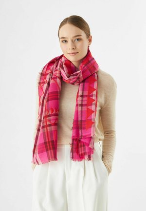 Jeune femme portant un pull beige et un pantalon blanc, avec une écharpe géométrique rose vif et rouge autour du cou.