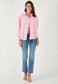 Light pink oversized shirt with long sleeves, button front, and soft fabric, paired with mid-rise flared blue jeans and multicolored sandals.