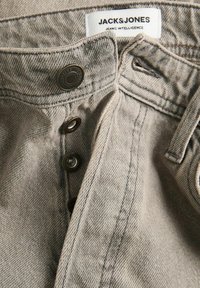 Közeli kép szürke farmernadrágról, nyitott gombos sliccel és jól látható fehér címkével, amelyen a „JACK & JONES JEANS INTELLIGENCE” felirat olvasható.